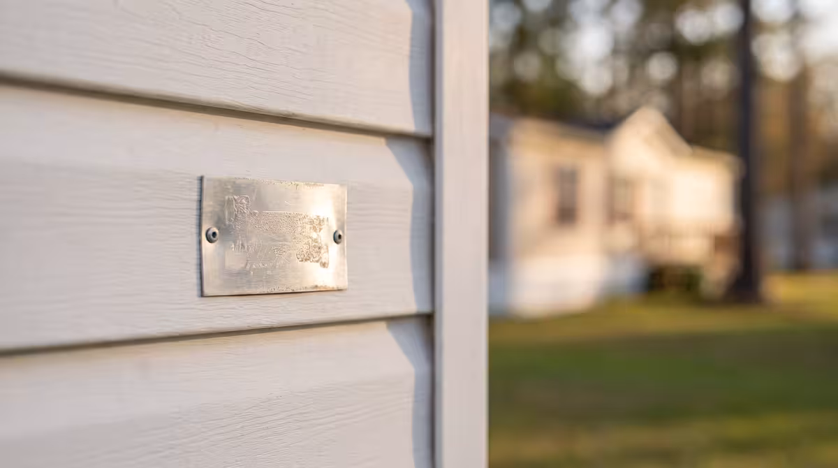 Weathered metal plate screwed to white mobile home siding, blurred house and trees in background.
