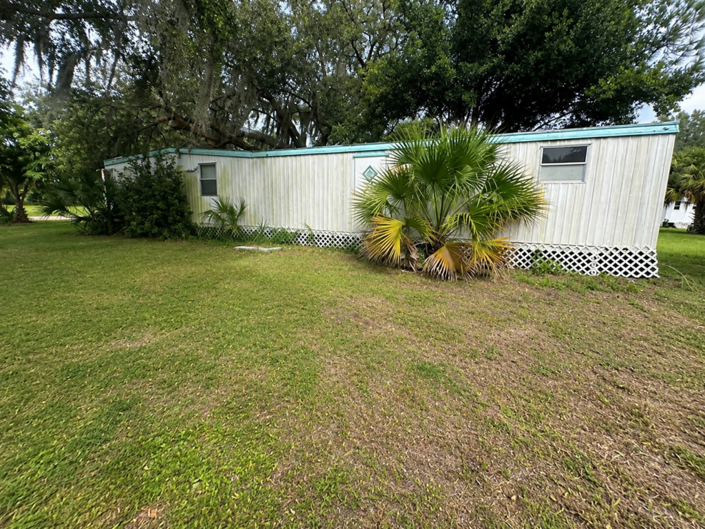 White mobile home with teal trim surrounded by grass, palm plants, and trees